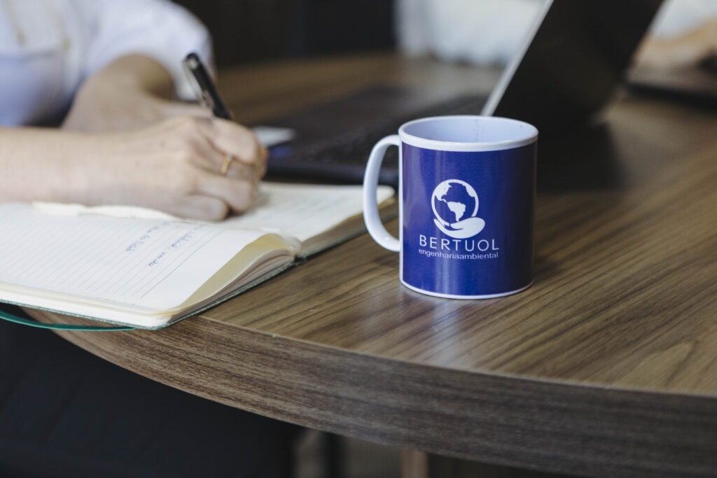 Pessoa fazendo anotações em um caderno ao lado de um notebook, com uma caneca da Bertuol Engenharia Ambiental sobre a mesa, representando ambiente de trabalho técnico e planejamento ambiental.
