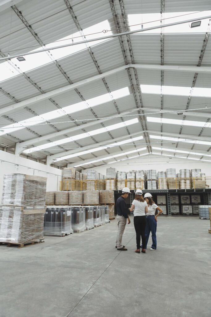 Galpão industrial com pallets de produtos armazenados e embalados, recipientes organizados em áreas específicas e três profissionais utilizando capacetes de segurança durante uma inspeção técnica no local.